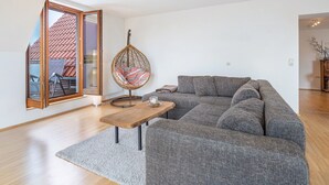 Uma sala de estar moderna com um sofá cinzento, uma mesa de centro de madeira, uma cadeira suspensa e uma grande janela com vista para um telhado de telhas vermelhas.