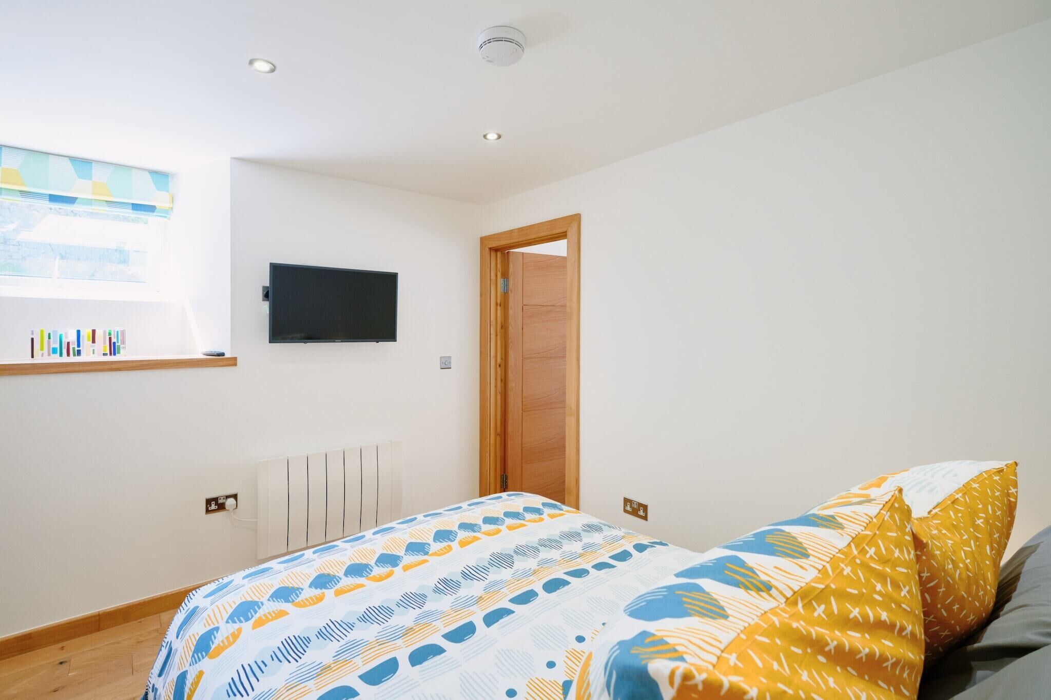 A bedroom with a bed, a TV mounted on the wall, a wooden door, and a window with blinds.