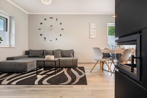 Un salon moderne avec un canapé d’angle, une horloge murale, un coin repas avec des chaises et une cuisine équipée d’un four encastré.
