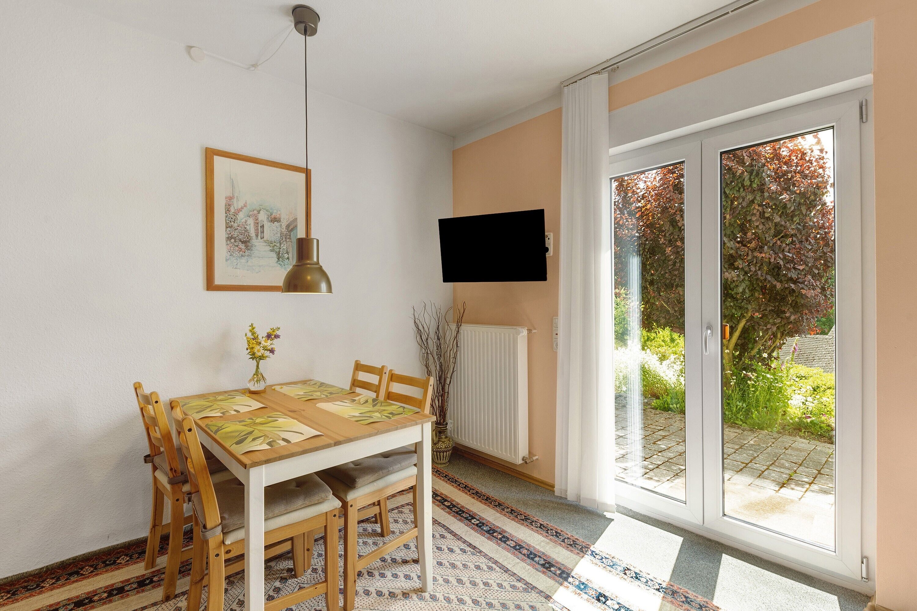 A dining area with a table and chairs, a painting on the wall, a television mounted above the table, and a sliding glass door leading to an outdoor area.