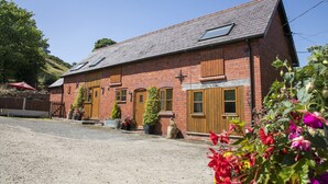 A red brick building with a sloped roof, two windows with shutters, and a wooden door. - Cottage in Dee Valley near Castell Dinas Bran (Llangollen)