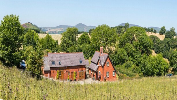 Une maison en briques rouges, dotée d’un toit en pente, entourée d’une végétation luxuriante et avec des collines en arrière-plan.