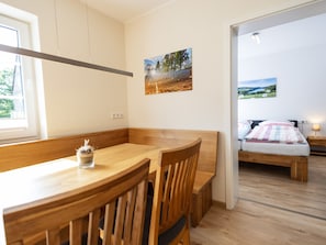 A room with a wooden dining table, chairs, a bed with a view, and a painting on the wall.
