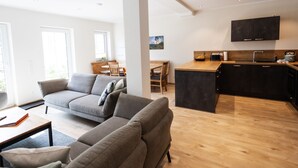 Een moderne woonkamer met een bank, een eethoek met een tafel en stoelen, een keuken met een spoelbak en een tv die boven het aanrecht is bevestigd.