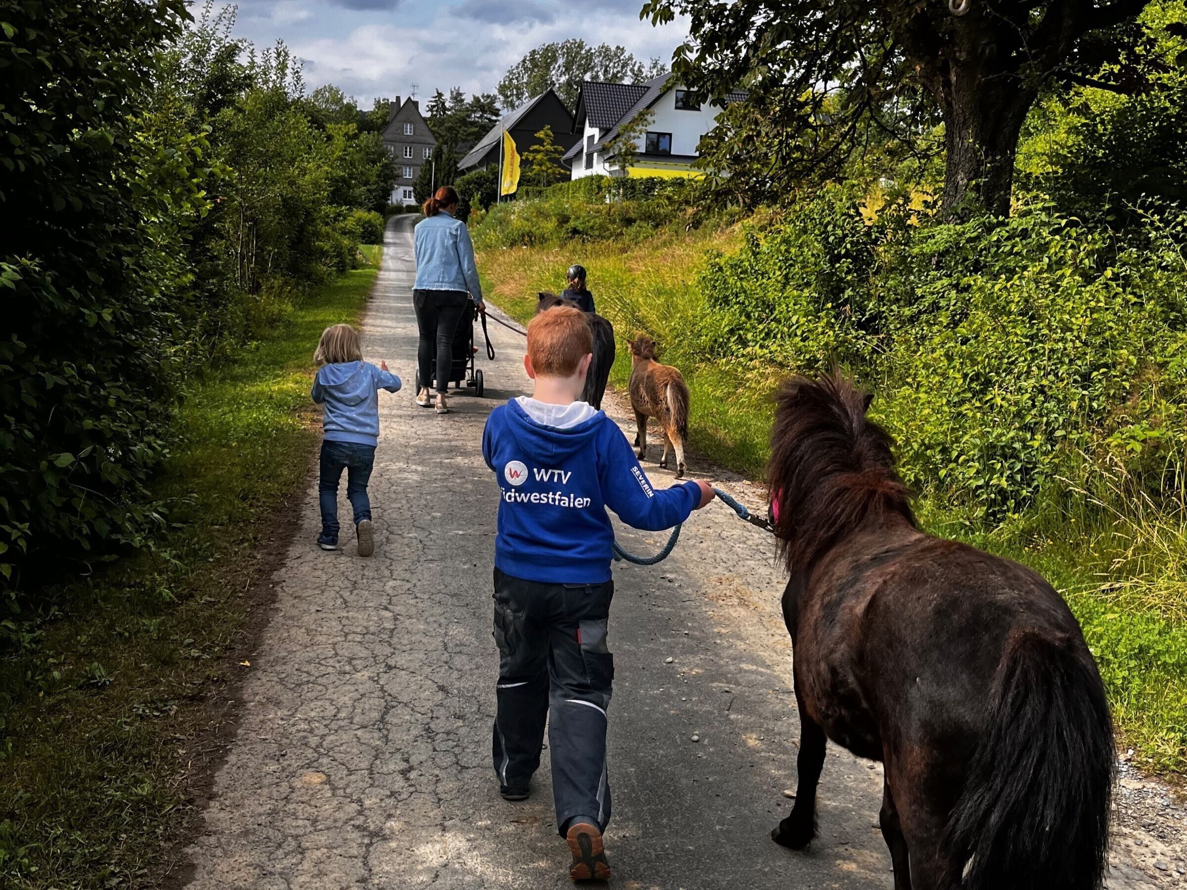 En stig med grönska på båda sidor, en person som skjuter en barnvagn och ett barn som går bredvid en ponny.