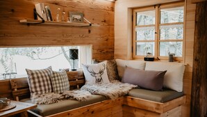 Interior acogedor de una cabaña con un banco de madera, una ventana con vistas a los árboles y un estante con libros y objetos decorativos.