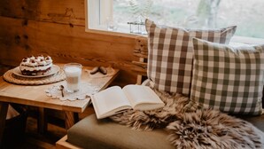 Interior acogedor de una cabaña con una mesa de madera, un pastel, un vaso de leche, un libro y almohadones a cuadros.