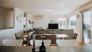 A modern living room with a grey sofa, a flat-screen TV, and a dining area with white chairs and a marble table.