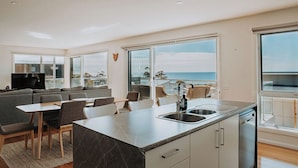 A modern living room with a sofa, dining table, and chairs, a kitchen with a sink and cabinets, and large windows offering a view of the sea.