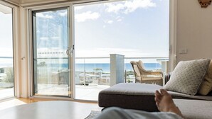 A room with a sofa and a table, a sliding glass door leading to a balcony with a view of the sea and buildings.