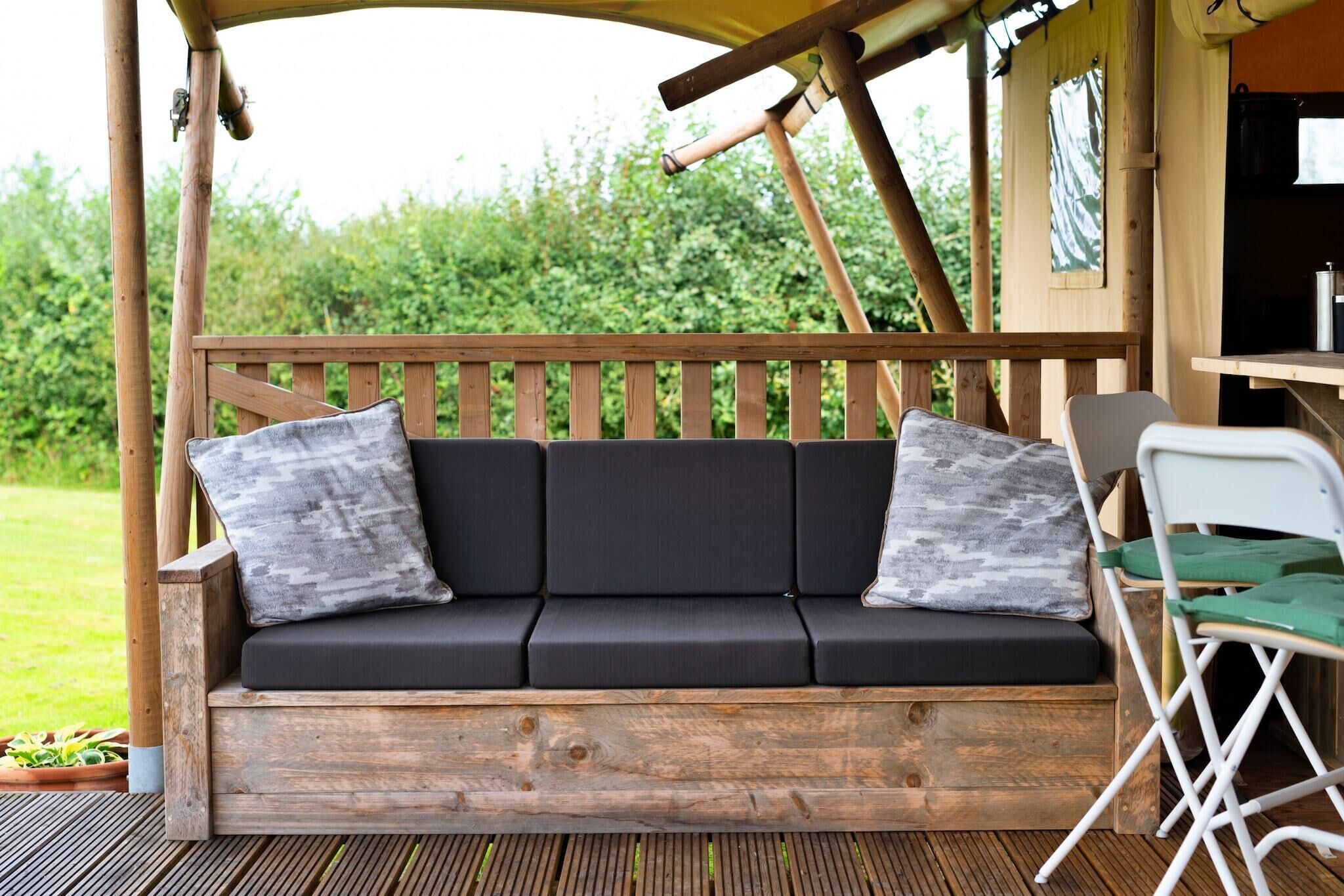 Een houten terras met een zwarte bank en twee kussens, een witte opklapbare stoel, en een kleine tafel met een waterkoker.