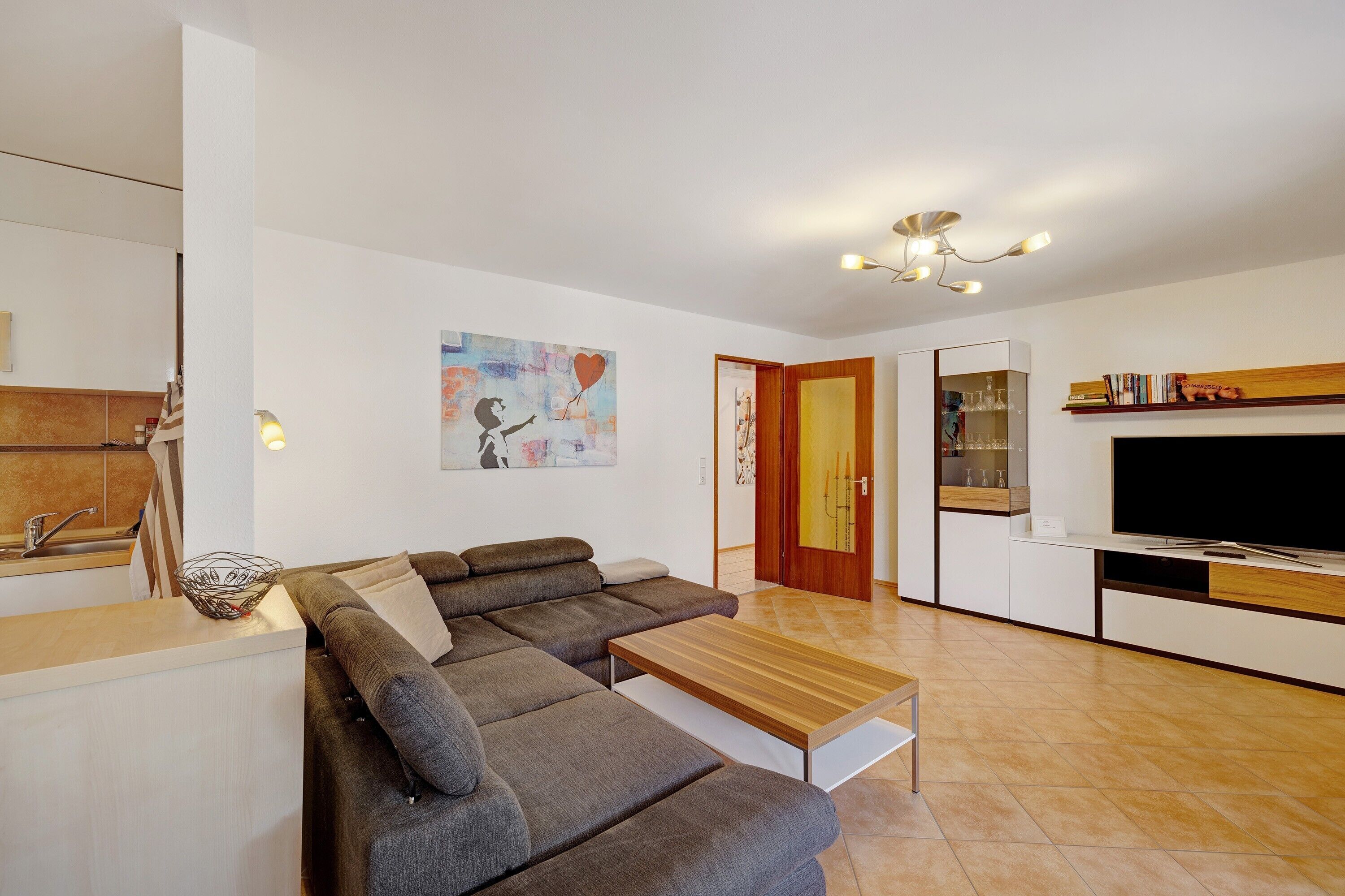 A modern living room with a sectional sofa, a wooden coffee table, a flat-screen TV, and a kitchen area with a countertop and cabinets.