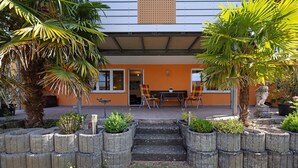 A modern building with a patio area featuring a table and chairs, surrounded by concrete planters and palm trees.