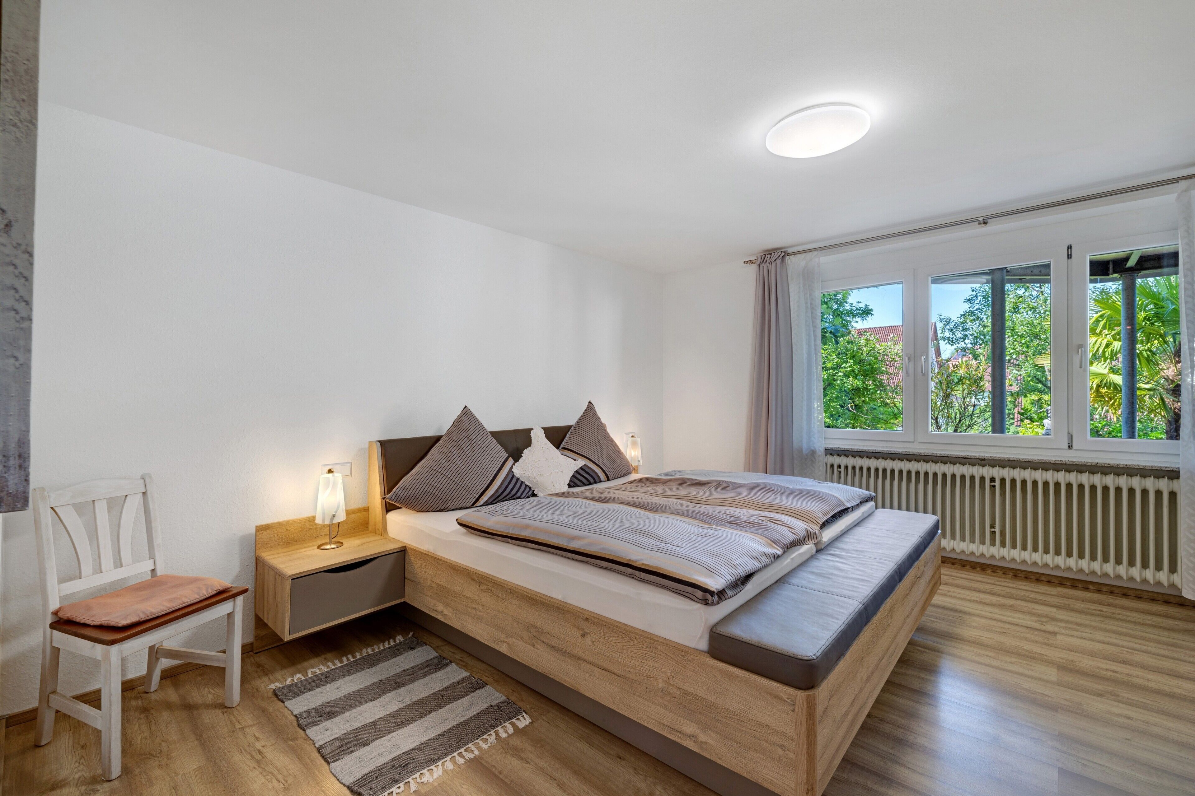 A bedroom with a bed, bedside table, chair, and a view of greenery through the window.