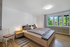 A bedroom with a bed, bedside table, chair, and a view of greenery through the window.
