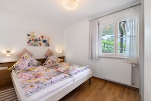 A bedroom with a bed, two bedside tables, a wall-mounted light, a window with curtains, and a radiator. - Apartment 'Weinberg' with Balcony (Meersburg)