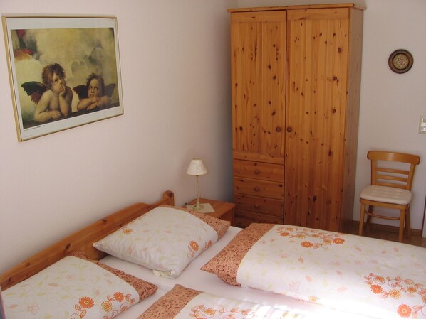 A bedroom with a wooden wardrobe, a bed with floral bedding, a chair, and a painting on the wall.