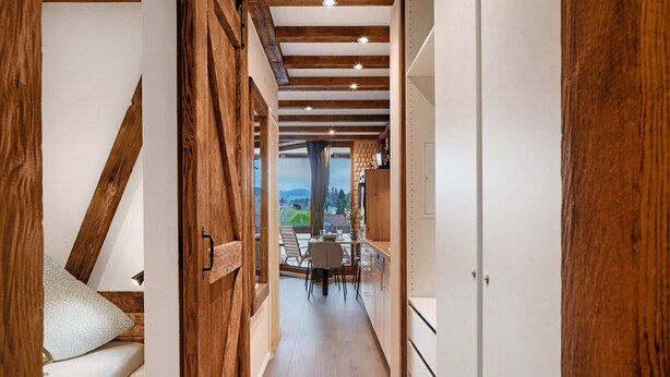 Un couloir étroit avec des poutres en bois, un lit avec un oreiller et un coin repas avec une table et des chaises.