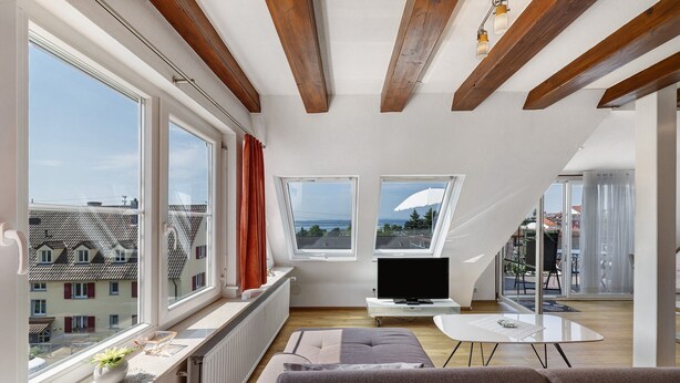 A modern living room with large windows, a flat-screen TV, a sofa, and a dining table. - Apartment 'Seeblick' with balcony, mountain and lake view (Meersburg)