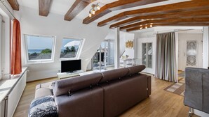 A modern living room with a brown sofa, wooden beams, large windows, and a television. - Apartment 'Seeblick' with balcony, mountain and lake view (Meersburg)
