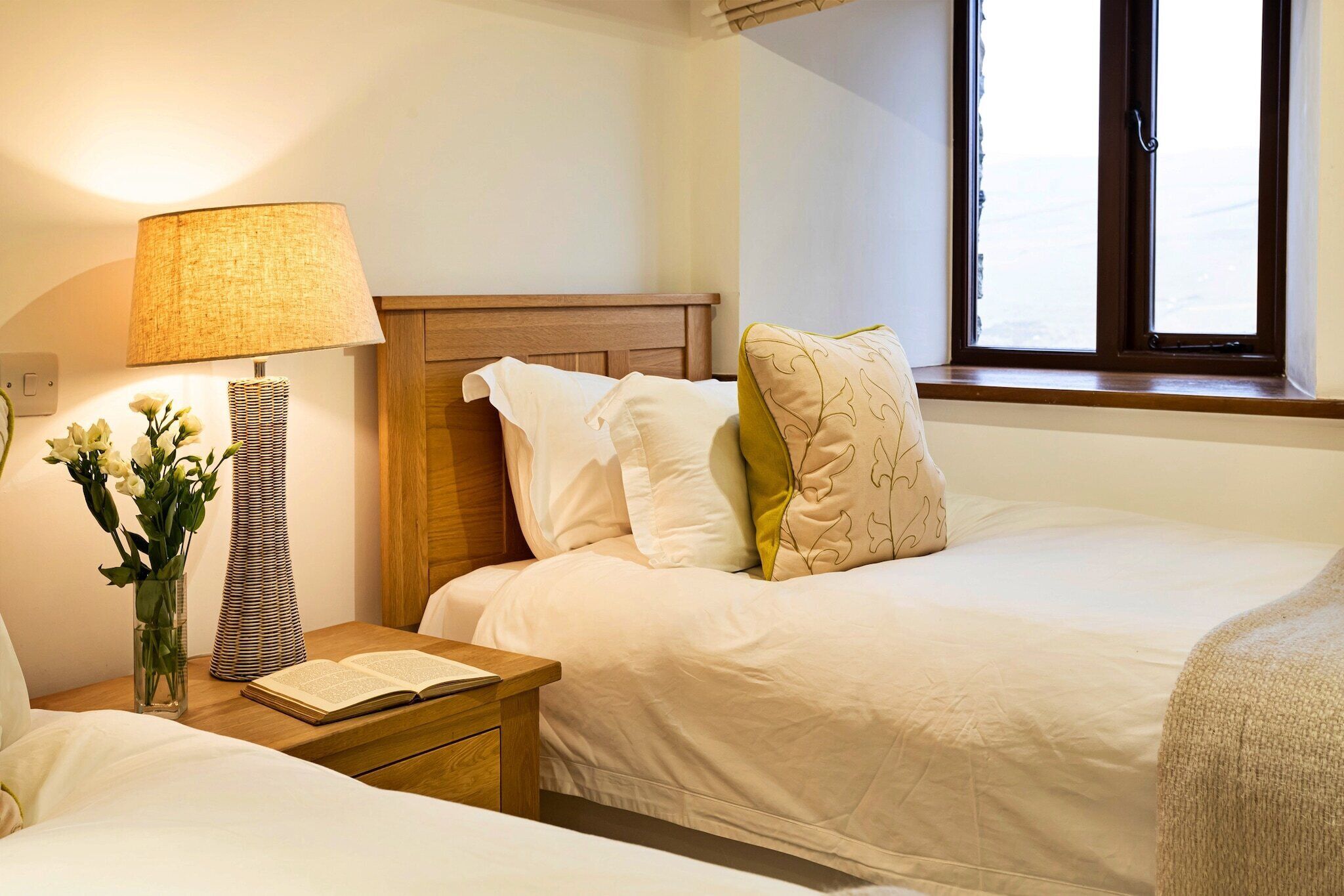 A hotel room with two beds, a nightstand with a lamp, a vase with flowers, and a window with a view.