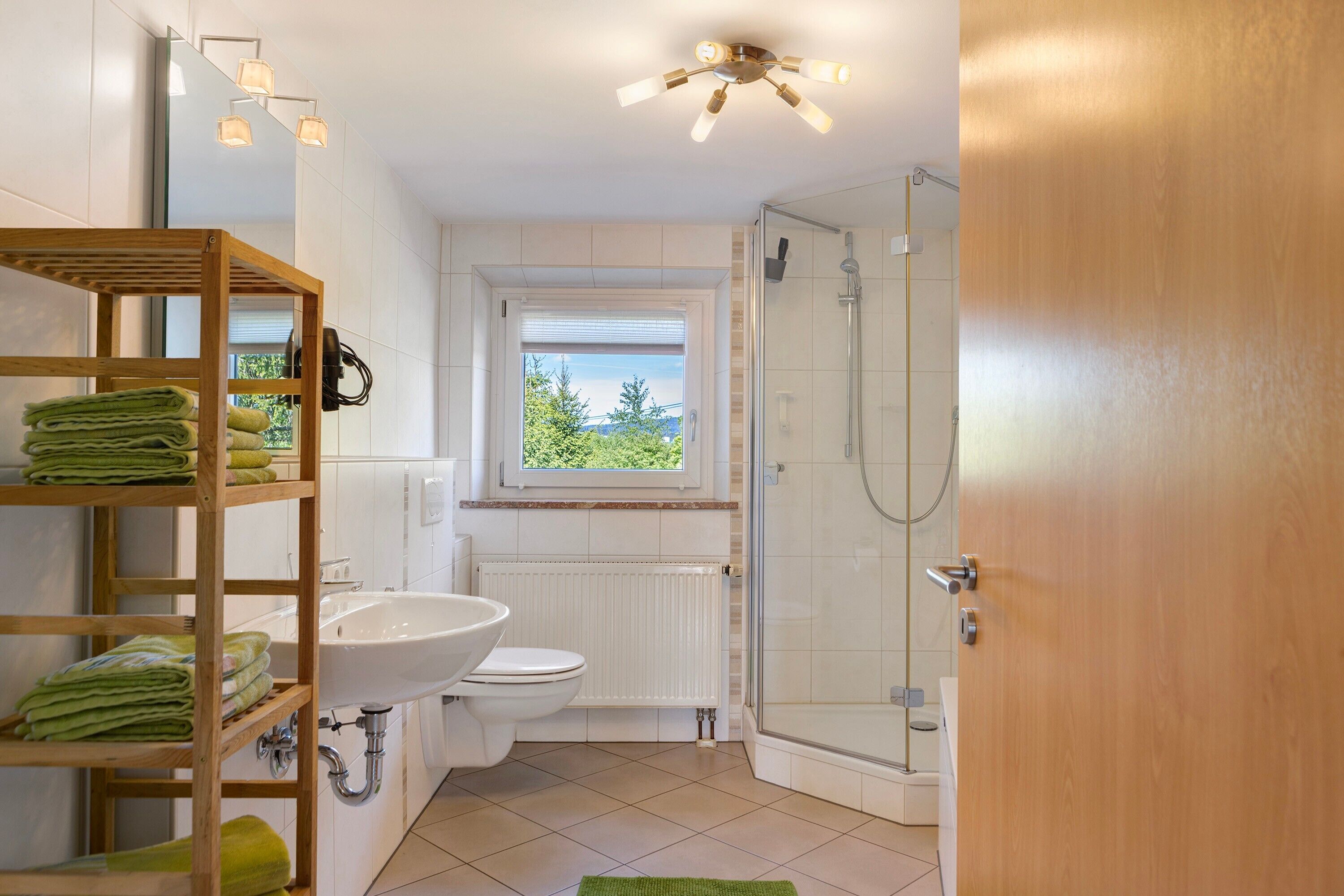 A bathroom with a toilet, sink, shower, and a wooden shelf with towels.