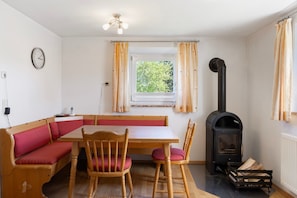 Una habitación con una mesa de madera, un banco acolchado rojo, dos sillas de madera, un estufa de leña negra, una ventana con cortinas y un reloj de pared.