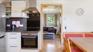 A compact kitchen with a stove, oven, and modern cabinetry. There is a dining area with a wooden table and chairs, and a sofa in the background. - Holiday Home 'Ferienhaus Kreuzbergblick' with Mountain View, Private Terrace and Wi-Fi (Hohenau)