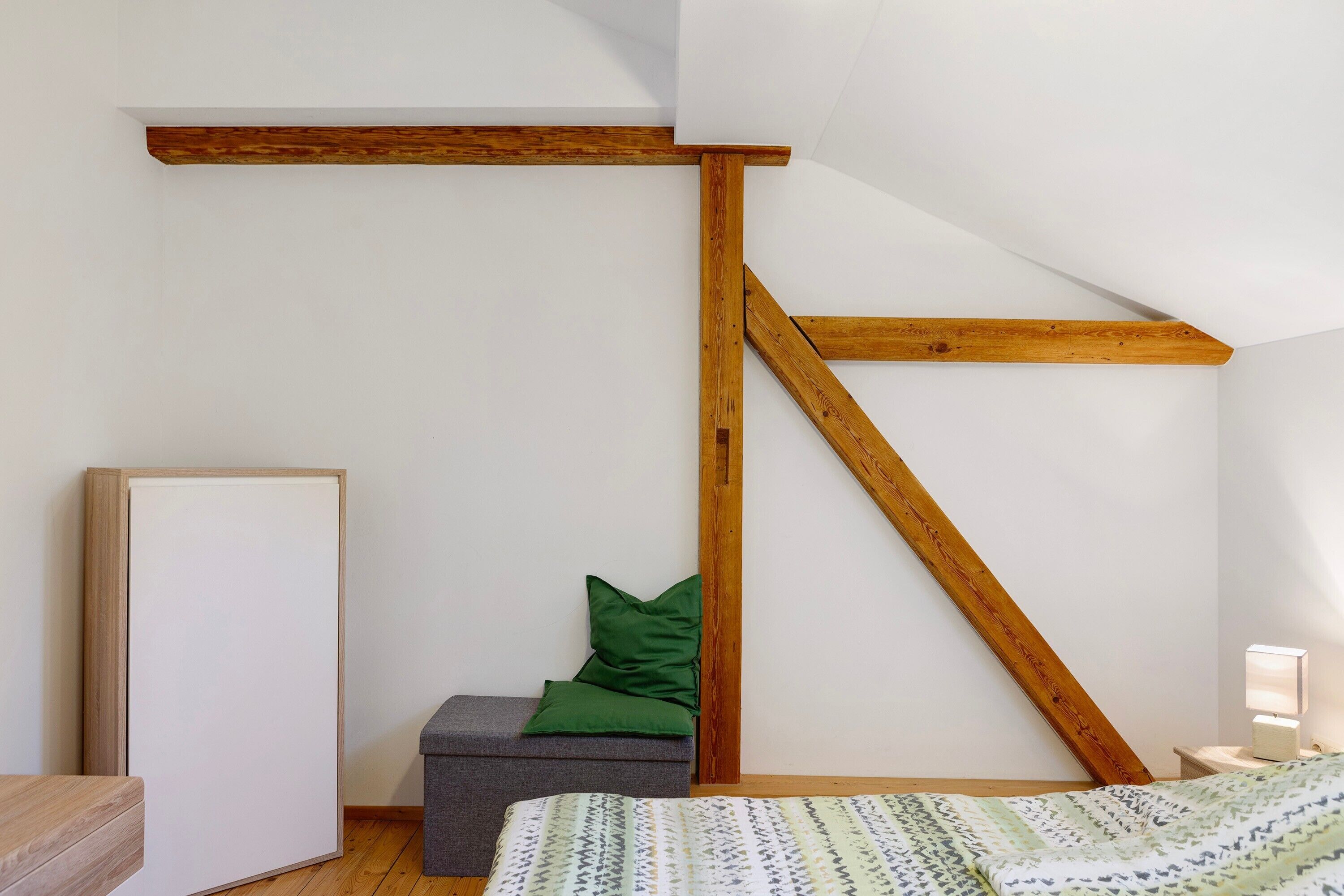 Ein Zimmer mit schrägem Dach, Holzbalken, einem Bett mit gemustertem Überwurf, einer Bank mit grünen Kissen und einem Nachttisch mit Lampe.