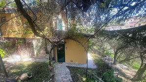 A stone pathway leading to a yellow building with a green door, surrounded by dense foliage and a metal fence.