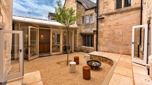 Une cour avec des murs en pierre, un patio fermé par des baies vitrées et un espace aménagé autour d’un foyer.
