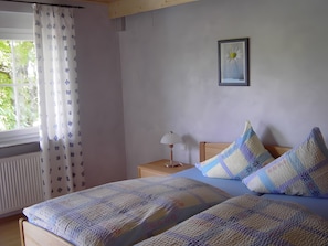 A bedroom with two beds, a bedside table with a lamp, a window with curtains, and a framed picture on the wall.
