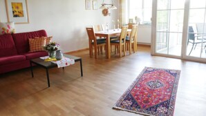 Una sala de estar con un sofá rojo, una mesa de centro con un jarrón, un comedor con sillas de madera y un ventanal grande.