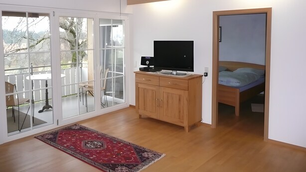 Una habitación moderna con techo de madera, un televisor sobre un mueble de madera, una cama con colchón azul y un amplio ventanal que da a un espacio exterior con sillas y una mesa.