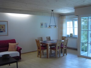 A dining area with a wooden table and chairs, a red sofa, and a window with a view of greenery.