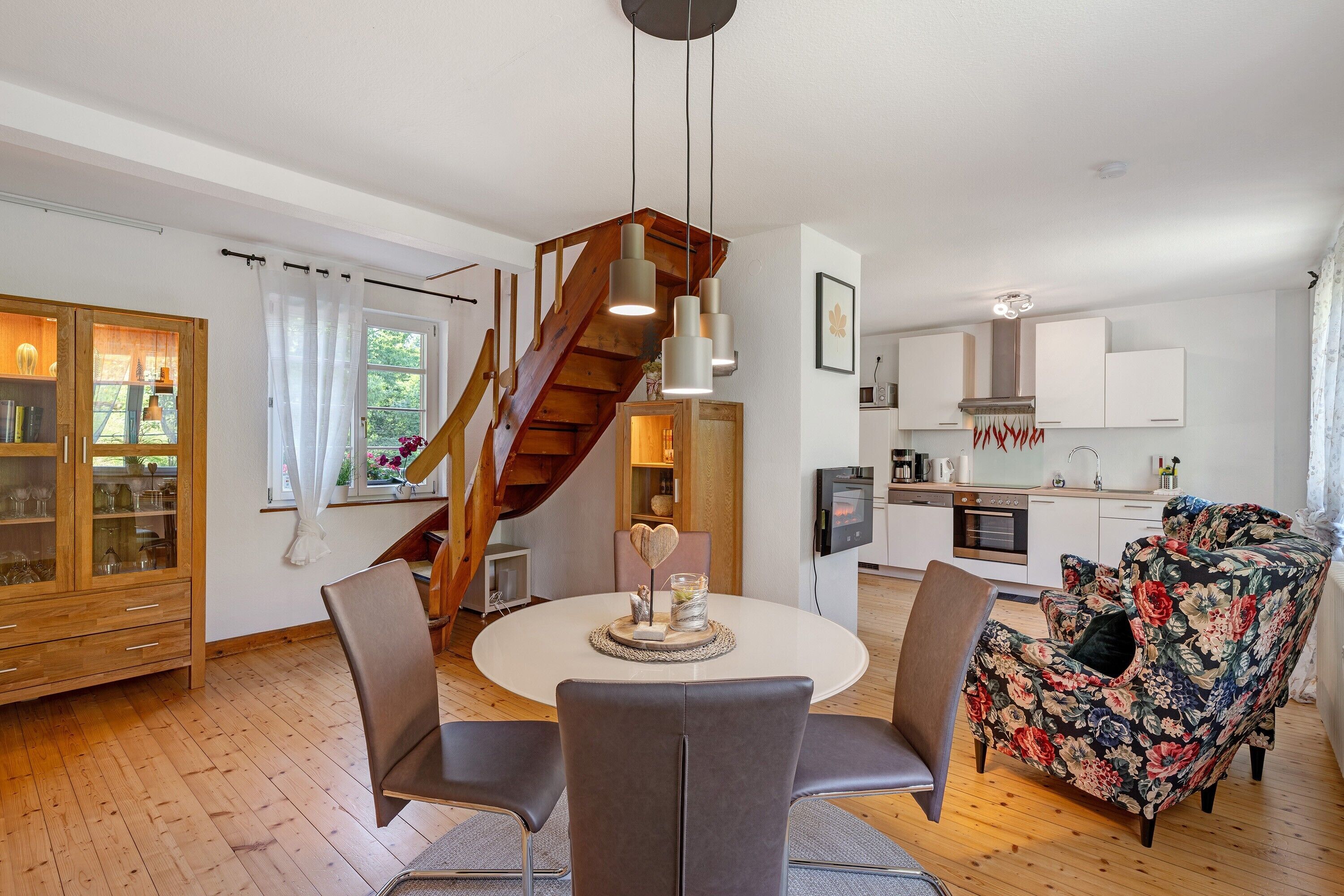 Eine moderne Küche mit weißen Schränken, einer Holztreppe, einem Essbereich mit rundem Tisch und Stühlen sowie einem Blumensofa.