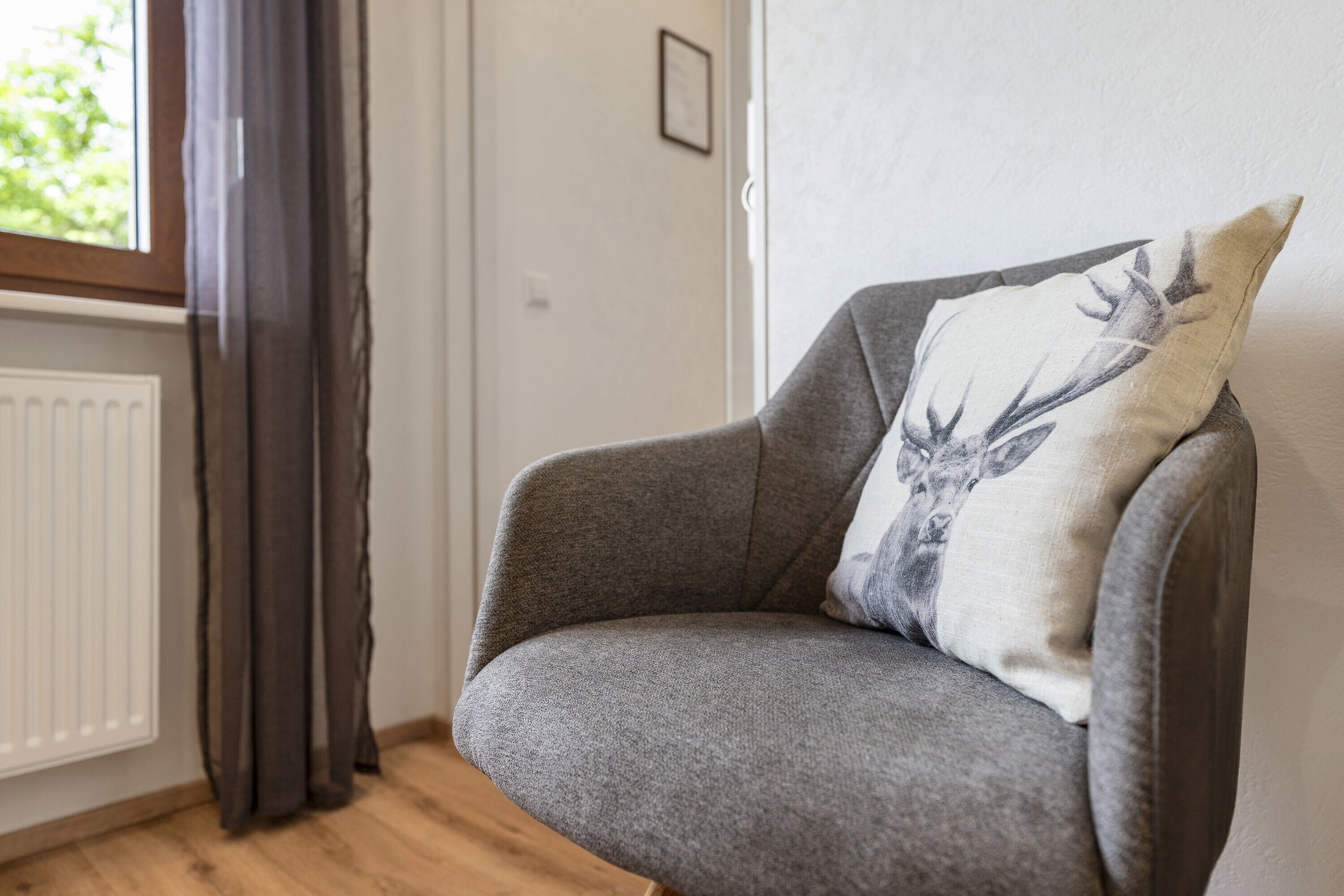 Ein grauer Sessel mit Kissen, verziert mit einem Hirschmuster, steht neben einem Fenster mit Vorhängen.