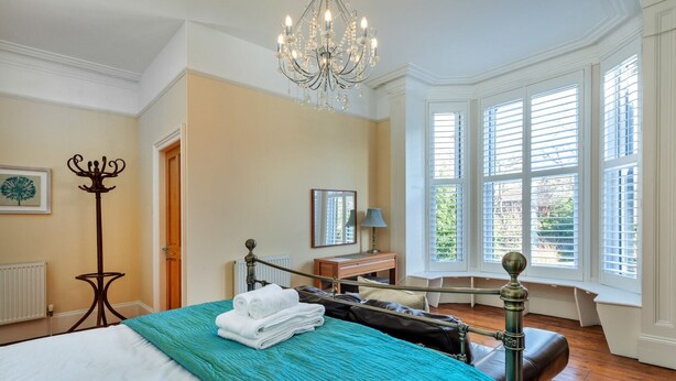 Un dormitorio bien iluminado con una cama grande, un candelabro, una ventana con persianas y vistas al exterior con vegetación.