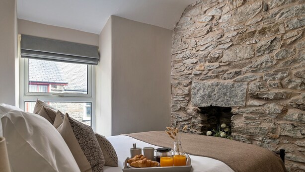 A bedroom with a stone wall, a bed with beige bedding, a tray with croissants, a cup, and orange juice.