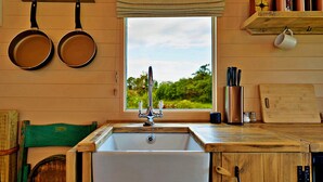A kitchen with a white sink, hanging pans, a window with a view, and wooden shelves with kitchen items.