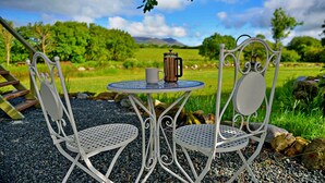 A metal table with two chairs, a coffee pot, and a cup on a gravel surface with a scenic view of trees and mountains in the background.