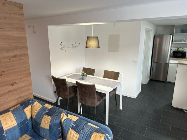 A modern living room with a sofa, dining table, chairs, and a kitchen area with a refrigerator and microwave.