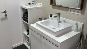 Un baño con lavamanos blanco, espejo y estantes con artículos de aseo.