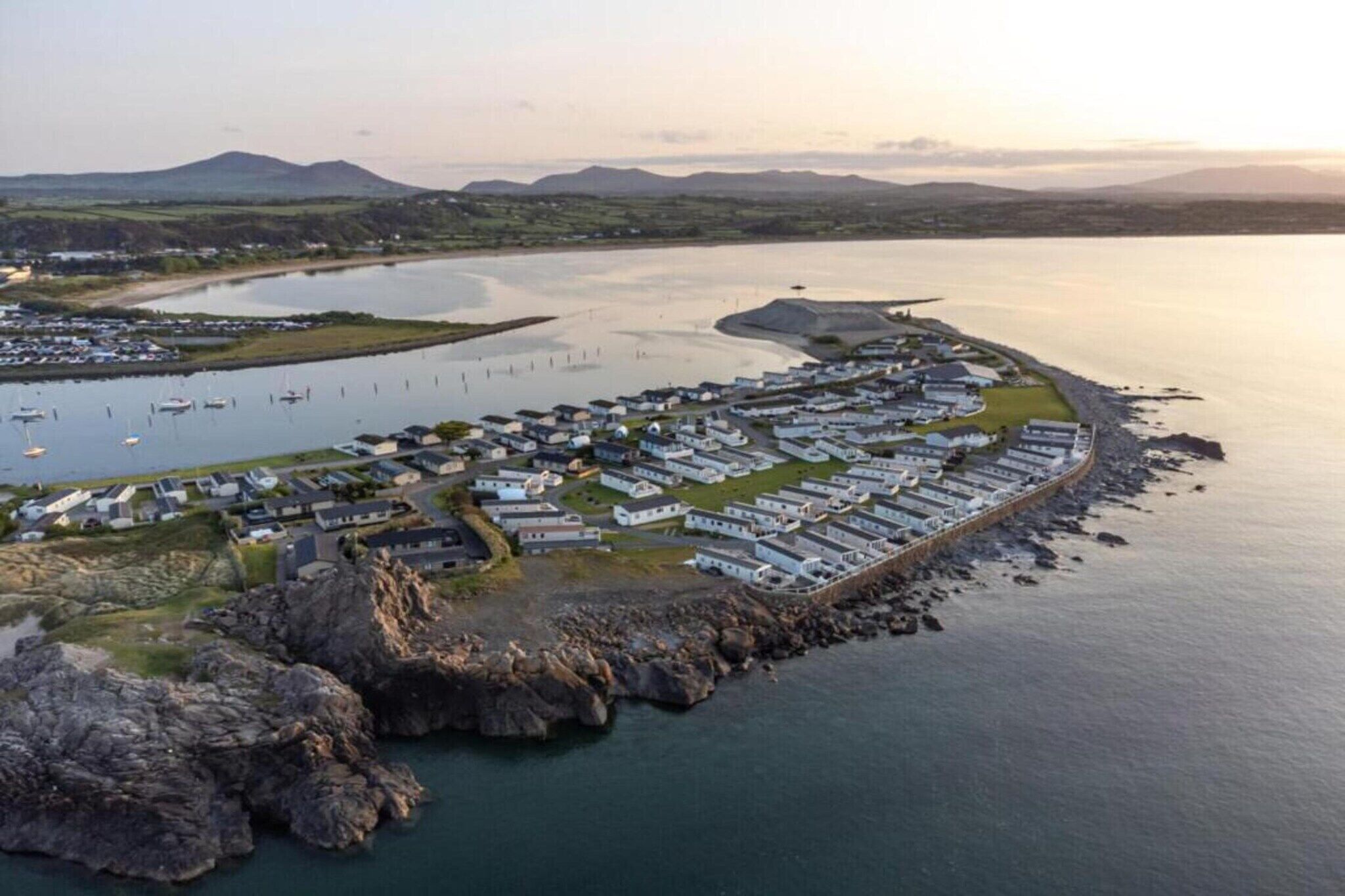 An aerial view of a coastal area with a row of white buildings along the shoreline, a marina with boats, and a rocky outcrop in the foreground.