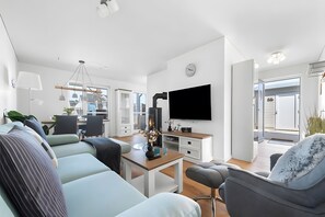 A modern living room with a sofa, armchair, and TV. There is a dining area with chairs and a table.