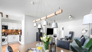 A modern kitchen with white cabinets, a dining area with a wooden table, and a living room with a sofa and a TV. - Holiday Apartment 'Maltes' with Sea View, Private Terrace and Wi-Fi (Kappeln)
