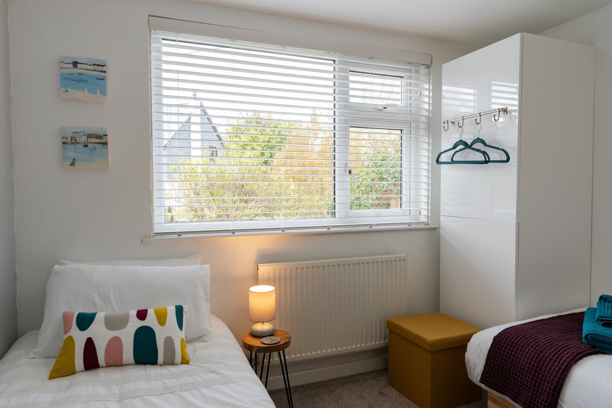 A bedroom with a bed, a bedside table with a lamp, a radiator, a window with blinds, and a wardrobe.