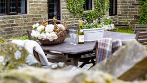 Une table de jardin rustique, dressée pour un repas en plein air, avec un panier de fleurs blanches, une bouteille de vin et un verre.