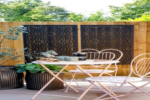Un patio al aire libre con una mesa y sillas, rodeado de macetas con plantas y una valla de madera decorativa.
