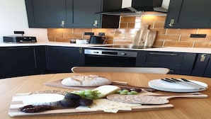 A kitchen with dark cabinets, a tiled backsplash, and a wooden table with a cheese board, bread, and cutlery.